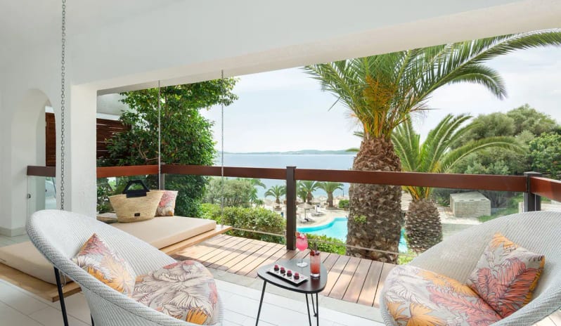 Bungalow Sea View High-end hotel balcony with a hanging daybed, tropical palm trees, cocktails on a side table, and a panoramic ocean view in Halkidiki at Eagles Palace by Kenwood Travel