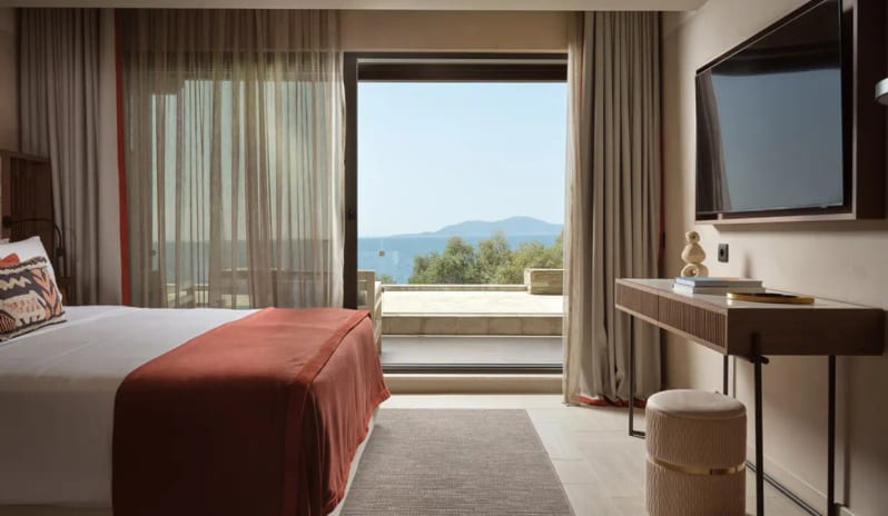 Deluxe Junior Suite Sea Front bedroom with warm wood accents, terracotta cushions, and a private balcony overlooking the Aegean Sea at Eagles Palace by Kenwood Travel