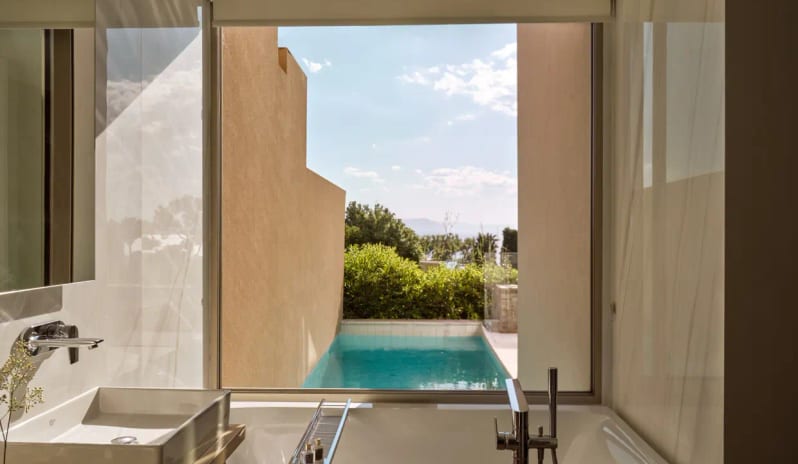 Junior Pool Villa Sea View bathroom interior featuring a large window that overlooks a private turquoise plunge pool and lush greenery at Eagles Resort in Halkidiki, Greece at Eagles Villas by Kenwood Travel