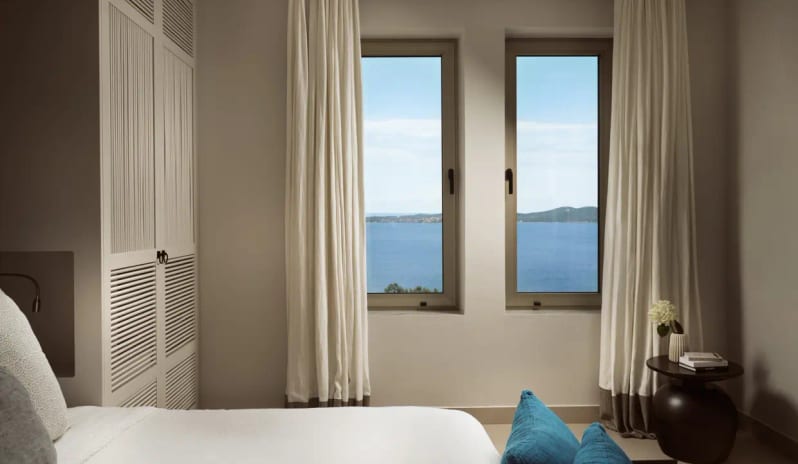 Residential 2 Bedroom Pool Villa Private Garden Sea View two large windows overlooking the Aegean Sea, white linens, neutral-toned curtains, and a modern side table with floral accents at Eagles Villas by Kenwood Travel