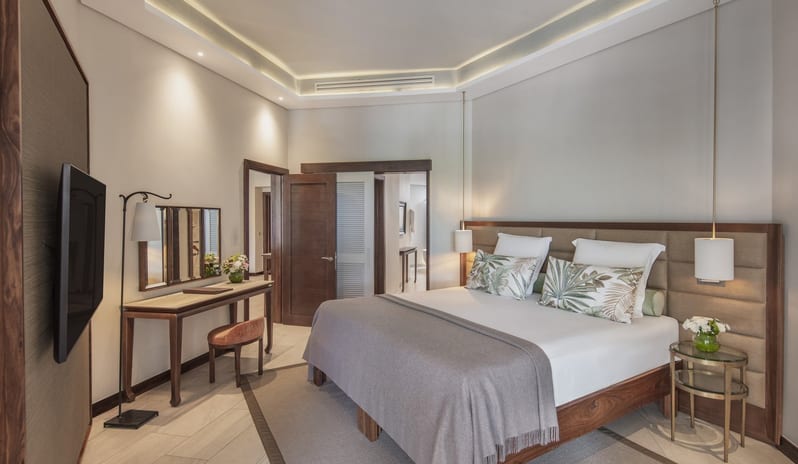 Penthouse Suite bedroom featuring a large wood-frame bed with neutral bedding, tropical print pillows, a sleek vanity desk with a tri-fold mirror, and recessed tray ceiling lighting at Royal Palm Beachcomber Luxury by Kenwood Travel