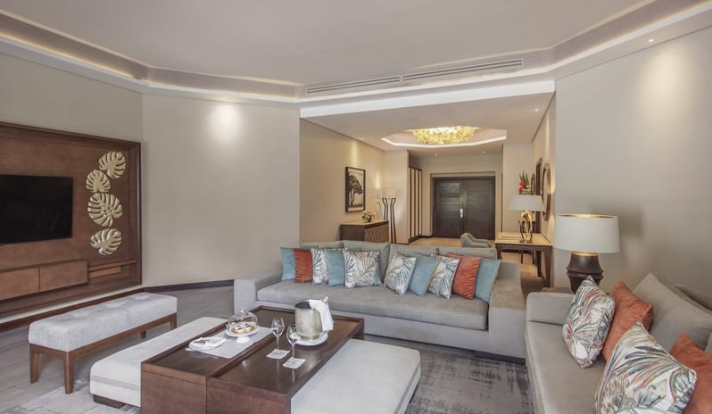 Penthouse Suite living room with a neutral-toned sectional sofa, wood-panelled TV wall with monstera leaf art, gold silk curtains, and large sliding glass doors opening to a tropical terrace at Royal Palm Beachcomber Luxury by Kenwood Travel