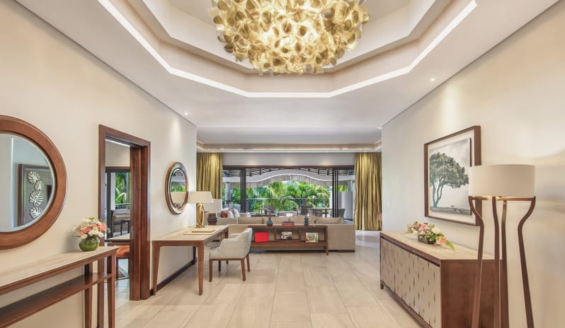 Penthouse Suite large, ornate golden chandelier in a recessed tray ceiling, wood-framed mirrors, console tables with floral arrangements, and a view leading out to a tropical balcon at Royal Palm Beachcomber Luxury by Kenwood Travel