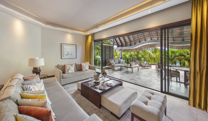 Penthouse Suite living room with a neutral-toned sectional sofa, wood-panelled TV wall with monstera leaf art, gold silk curtains, and large sliding glass doors opening to a tropical terrace at Royal Palm Beachcomber Luxury by Kenwood Travel