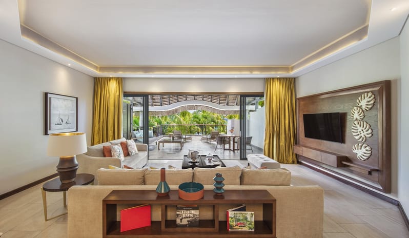 Penthouse Suite living room with a neutral-toned sectional sofa, wood-panelled TV wall with monstera leaf art, gold silk curtains, and large sliding glass doors opening to a tropical terrace at Royal Palm Beachcomber Luxury by Kenwood Travel