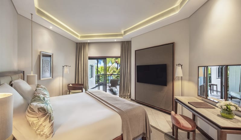 Penthouse Suite bedroom featuring a large wood-frame bed with neutral bedding, tropical print pillows, a sleek vanity desk with a tri-fold mirror, and recessed tray ceiling lighting at Royal Palm Beachcomber Luxury by Kenwood Travel