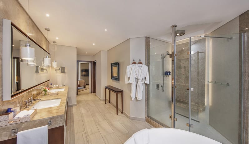 Penthouse Suite bathroom with a marble double vanity, large glass-enclosed walk-in shower, a freestanding soaking tub, and neutral stone tiling throughout at Royal Palm Beachcomber Luxury by Kenwood Travel