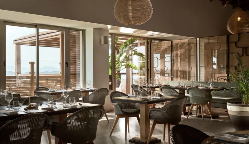 Lofos Restaurant wicker chairs, modern wooden tables, and large glass doors overlooking the sea at Eagles Villas by Kenwood Travel