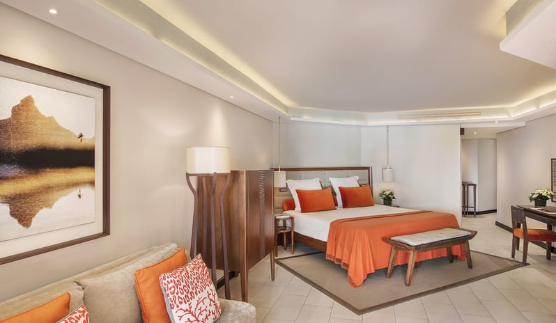 Junior Suite Spacious luxury hotel suite interior featuring a king bed with orange linens, a cozy sofa with decorative pillows, and a dedicated workspace desk at Royal Palm Beachcomber Luxury by Kenwood Travel