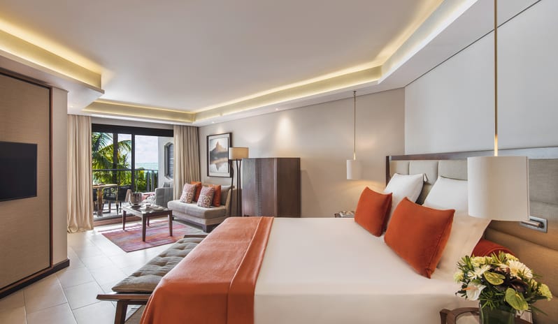 Junior Suite king-sized bed with orange pillows, a seating area, and a private balcony overlooking palm trees and the ocean at Royal Palm Beachcomber Luxury by Kenwood Travel