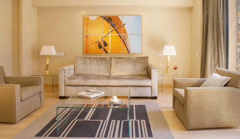 The Collection Premium One Bedroom Suite Sea View living room with a beige velvet sofa, two armchairs, a glass coffee table, and large-scale maritime wall art under recessed warm lighting at Daios Cove by Kenwood Travel