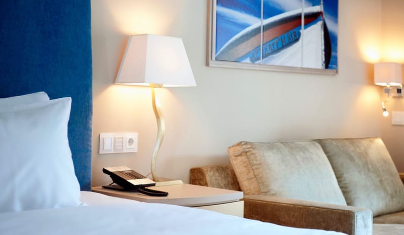 The Collection One bedroom Suite Sea View Private Pool white bed with a blue headboard, a bedside table with a curved gold lamp and telephone, and a beige velvet sofa next to coastal wall art at Daios Cove by Kenwood Travel