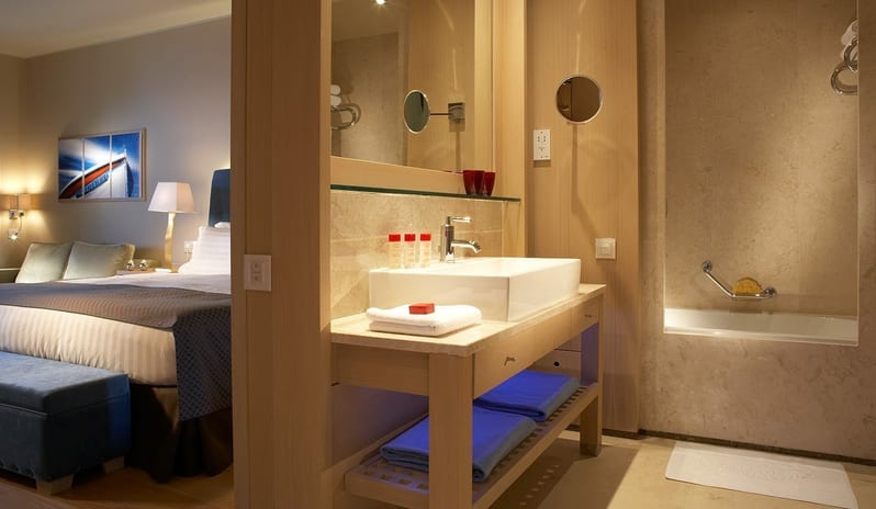 The Collection Premium One Bedroom Suite Sea View bathroom with a vessel sink and marble bathtub adjacent to a bedroom with a king-size bed and blue accents at Daios Cove by Kenwood Travel