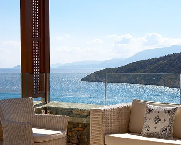 The Collection Premium One Bedroom Suite Sea View terrace with white wicker furniture under a wooden pergola, overlooking a blue bay and coastal mountains at Daios Cove by Kenwood Travel