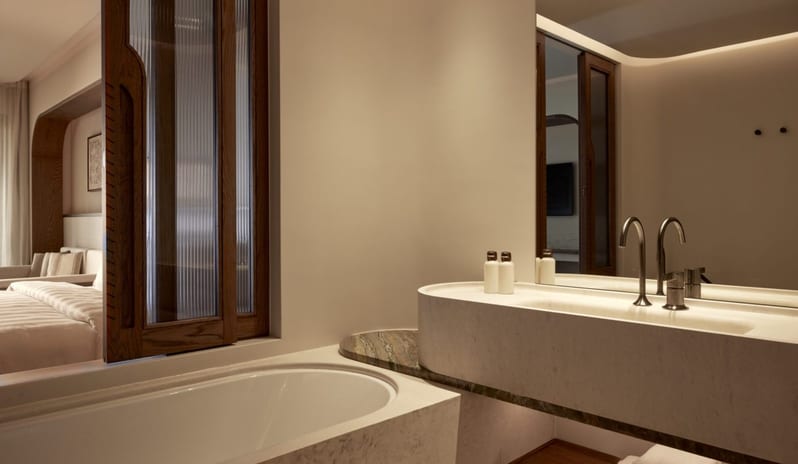 The Collection Junior Suite Sea View Private Pool bathroom featuring a deep soaking tub, a carved stone double vanity with sleek black faucets, and a reeded glass window looking into the bedroom suite at Daios Cove by Kenwood Travel