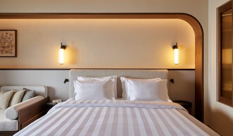 The Collection Junior Suite Sea View Private Pool bedroom featuring a plush bed with striped linens, curved wood wall alcove, warm sconce lighting, and a neutral daybed at Daios Cove by Kenwood Travel