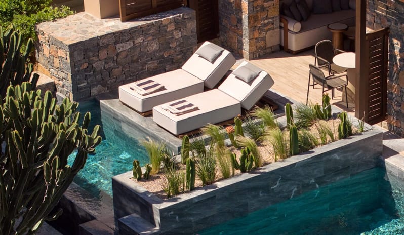 Tterrace featuring a plunge pool, two modern sun loungers on a floating platform, stone walls, and desert landscaping with cacti at Daios Cove by Kenwood Travel