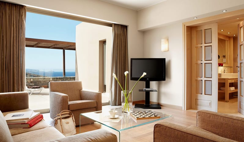 The Collection One bedroom Suite Sea View Private Pool living room with beige modern furniture, a glass coffee table with a chessboard, and large sliding doors opening to a private stone terrace with a Mediterranean sea view at Daios Cove by Kenwood Travel