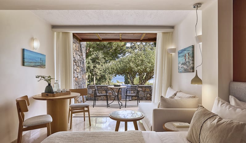 Classic Double Room Limited Sea View white interior, a private stone-walled terrace with outdoor seating, and a limited view of the sea through lush green trees at St. Nicolas Bay Resort by Kenwood Travel