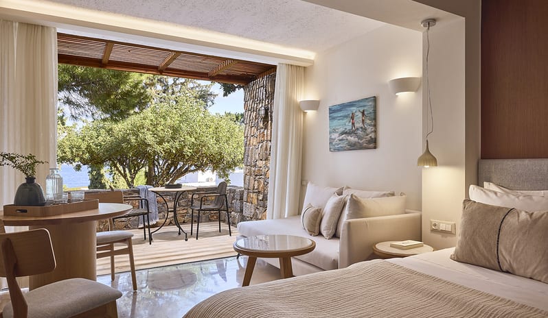 Classic Double Room Limited Sea View white interior, a private stone-walled terrace with outdoor seating, and a limited view of the sea through lush green trees at St. Nicolas Bay Resort by Kenwood Travel