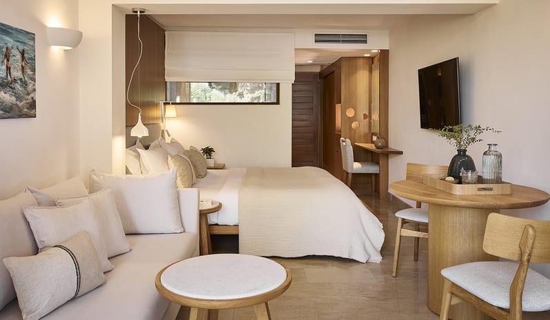 Classic Double Room Limited Sea View light-colored sofa, wooden dining table, and a double bed with neutral linens, highlighting the warm wood accents and minimalist design at St. Nicolas Bay Resort by Kenwood Travel