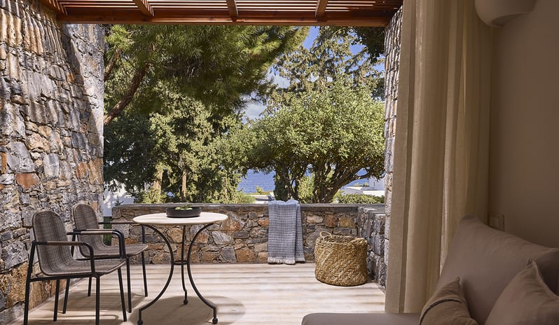 Classic Double Room Limited Sea View private hotel terrace with rustic stone walls and a wooden pergola, featuring a bistro table and chairs overlooking lush green trees with a partial view of the blue sea at St. Nicolas Bay Resort by Kenwood Travel