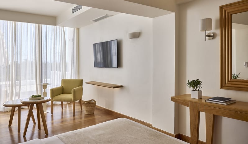 Classic Junior Suite Sea View warm wood flooring, a yellow armchair, a wall-mounted flat-screen TV, and a large window with sheer white curtainsat St. Nicolas Bay Resort by Kenwood Travel