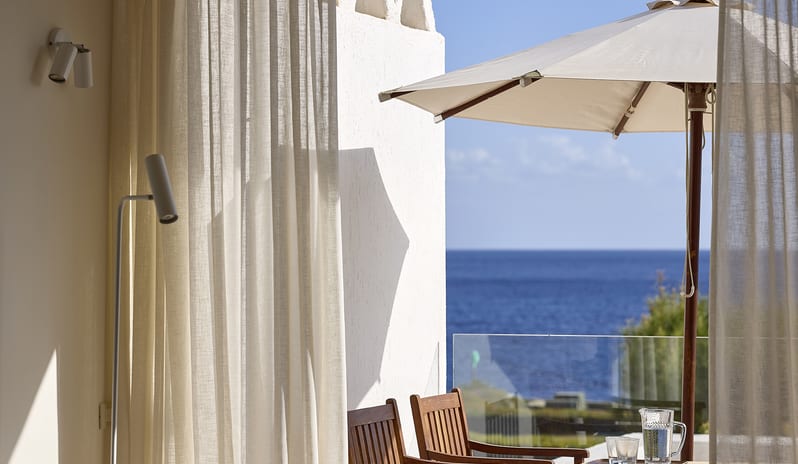 Classic Junior Suite Sea View wbalcony overlooking the deep blue sea, featuring wooden patio chairs, a white sun umbrella, and sheer white curtains blowing in the breeze at St. Nicolas Bay Resort by Kenwood Travel