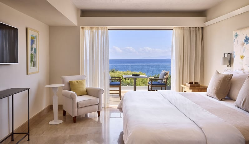 Classic Suite One Bedroom Sea View bedroom opening onto a sunlit private balcony with a white table and blue-cushioned chairs, overlooking a bright blue sea and distant island at St. Nicolas Bay Resort by Kenwood Travel