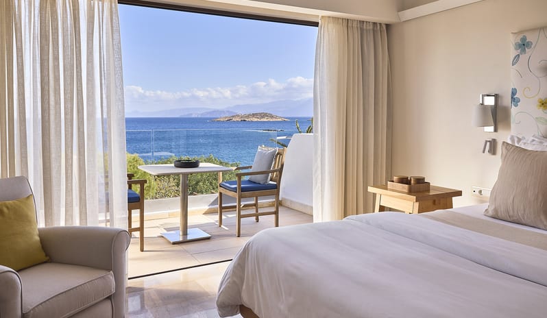 Classic Suite One Bedroom Sea View bedroom opening onto a sunlit private balcony with a white table and blue-cushioned chairs, overlooking a bright blue sea and distant island at St. Nicolas Bay Resort by Kenwood Travel