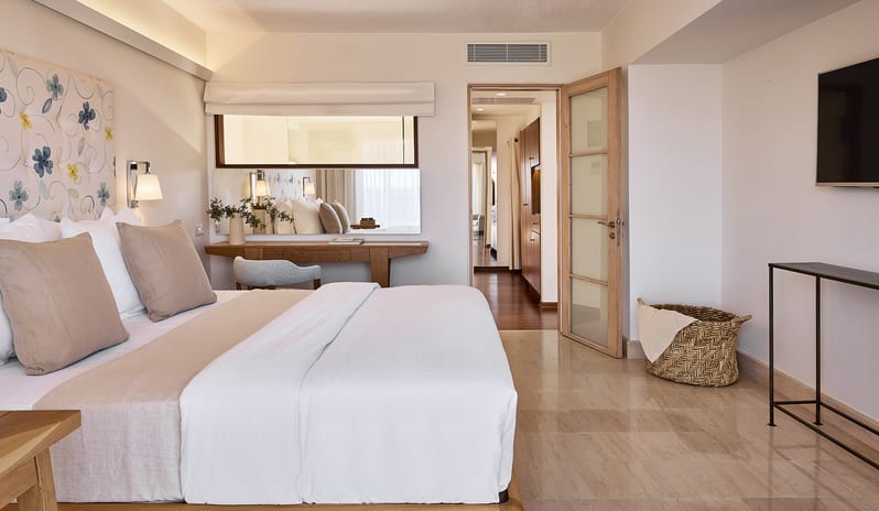 Classic Suite One Bedroom Sea View king-size bed with floral headboard, marble flooring, a wooden desk with a large mirror, and an open door leading to a hallway at St. Nicolas Bay Resort by Kenwood Travel