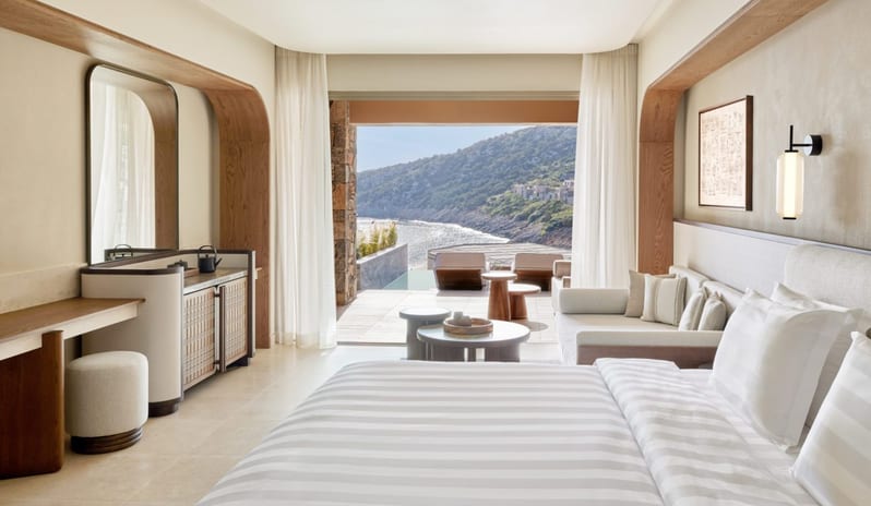 The Collection Two Bedroom Suite Sea View Private Pooll bedroom with a white striped bed, a minimalist desk and vanity area, and large glass doors opening to a scenic balcony overlooking the sea at Daios Cove by Kenwood Travel