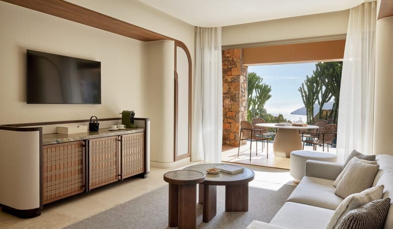 The Collection Two Bedroom Suite Sea View Private Pooll living room with a wall-mounted TV, marble-topped sideboard, and a sun-drenched terrace with an outdoor dining set overlooking the ocean at Daios Cove by Kenwood Travel