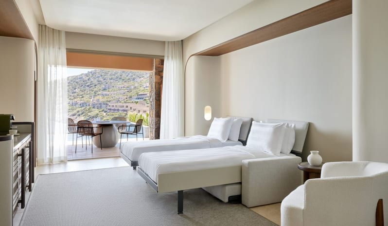 The Collection Two Bedroom Suite Sea View Private Pooll bedroom with two twin pull-out wall beds, a minimalist white armchair, and a balcony overlooking a coastal hillside village at Daios Cove by Kenwood Travel