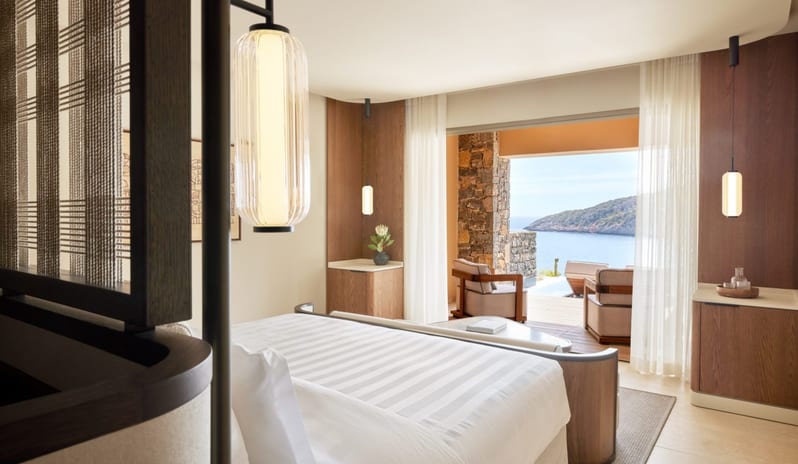 The Collection Two Bedroom Suite Sea View Private Pooll bedroom with an ocean view, featuring a white striped bed, minimalist wood furniture, and a private terrace with lounge chairs overlooking the sea at Daios Cove by Kenwood Travel