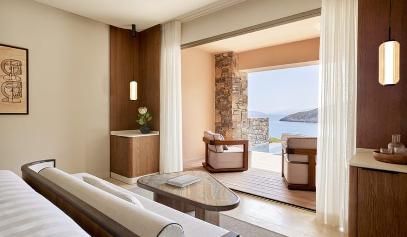 The Collection Two Bedroom Suite Sea View Private Poollbedroom suite lounge area with a designer sofa and marble table, looking out through an open doorway to a private wooden deck with armchairs and an ocean view at Daios Cove by Kenwood Travel