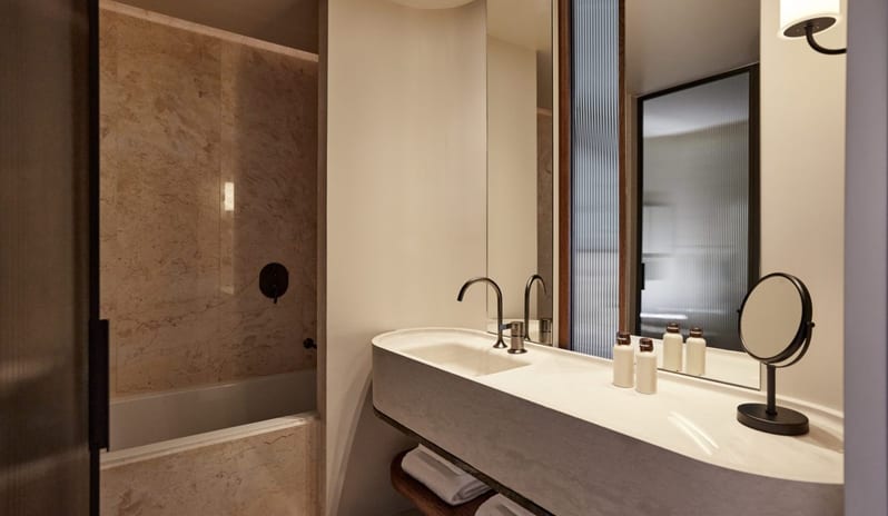 The Collection Junior Suite Sea View bathroom featuring a large marble-clad bathtub and an integrated stone vanity with a modern black faucet, fluted glass partitions, and a large mirror reflecting warm ambient lighting at Daios Cove by Kenwood Travel