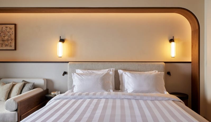 The Collection Junior Suite Sea View bedroom with a wood-framed bed nook, glowing wall lamps, and white striped bedding at Daios Cove by Kenwood Travel