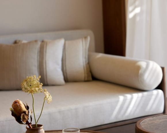The Collection Junior Suite Sea View brown stone coffee table with a small floral arrangement and a glass of water, set against a blurred cream-colored sofa with striped cushionsat Daios Cove by Kenwood Travel