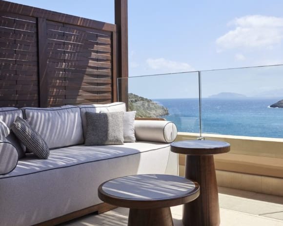 The Collection Junior Suite Sea View private terrace of a junior suite overlooking the blue ocean, featuring a large outdoor sofa with striped pillows, wooden pedestal side tables, and a shaded pergola at Daios Cove by Kenwood Travel