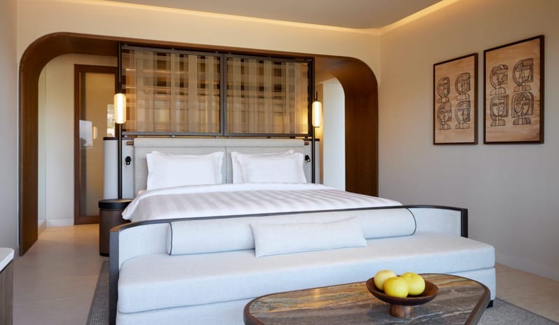 The Collection One bedroom Suite Sea View Private Pool king-size bed with a textured wood room divider, a curved gray settee, and a marble coffee table under warm cove lighting at Daios Cove by Kenwood Travel