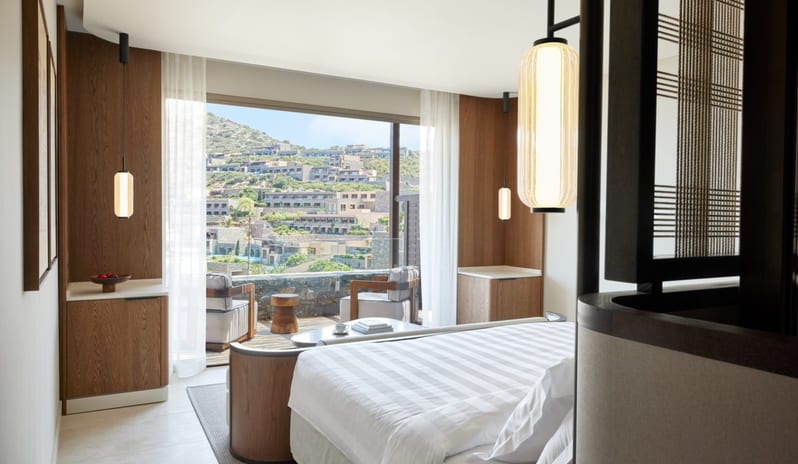 The Collection One bedroom Suite Sea View Private Pool king-size bed overlooking a private balcony with lounge chairs and a view of a coastal hillside development under a clear sky at Daios Cove by Kenwood Travel