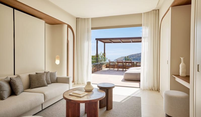 The Collection One bedroom Suite Sea View Private Pool outdoor terrace with a wooden dining table under a pergola, offering a panoramic view of the blue ocean and rugged coastal mountains at Daios Cove by Kenwood Travel