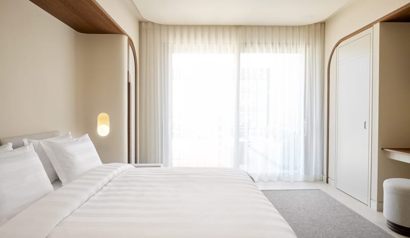 The Collection One bedroom Suite Sea View Private Pool living room featuring a fold-down wall bed with white striped linens, soft cove lighting, and floor-to-ceiling sheer white curtains filtering natural light at Daios Cove by Kenwood Travel