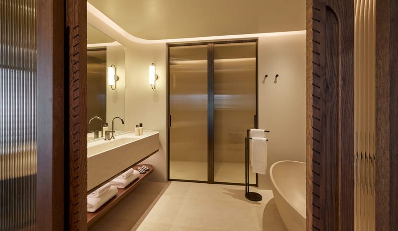 The Collection One bedroom Suite Sea View Private Pool bathroom featuring a large freestanding soaking tub, a long stone vanity with double faucets, and fluted glass shower doors under soft cove lighting at Daios Cove by Kenwood Travel