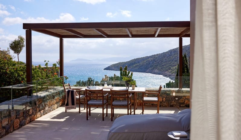 The Collection One bedroom Suite Sea View Private Pool terrace with a wooden dining table and chairs under a pergola, offering a panoramic view of a blue sea and rugged coastal mountains at Daios Cove by Kenwood Travel