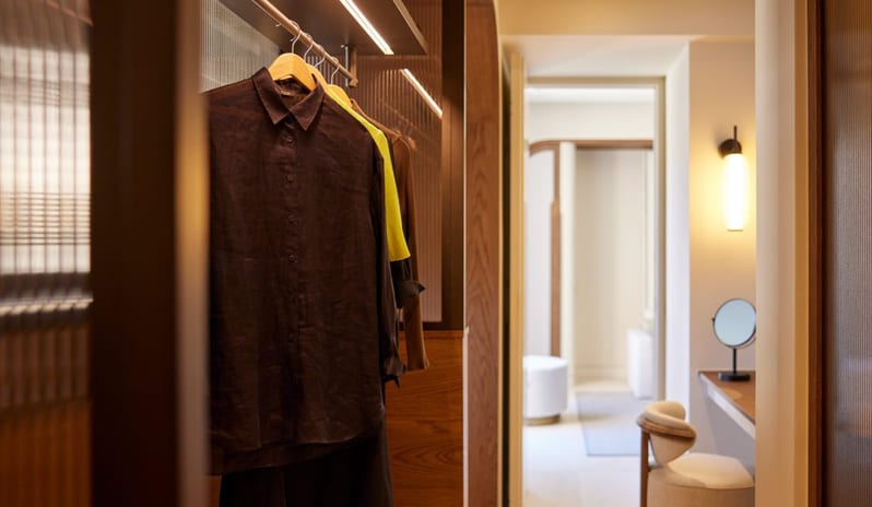 The Collection One bedroom Suite Sea View Private Pool luxury hotel bedroom featuring a textured wood room divider, a modern cylindrical pendant light, and an arched wood-lined entryway leading to a private vanity desk with a circular stool at Daios Cove by Kenwood Travel