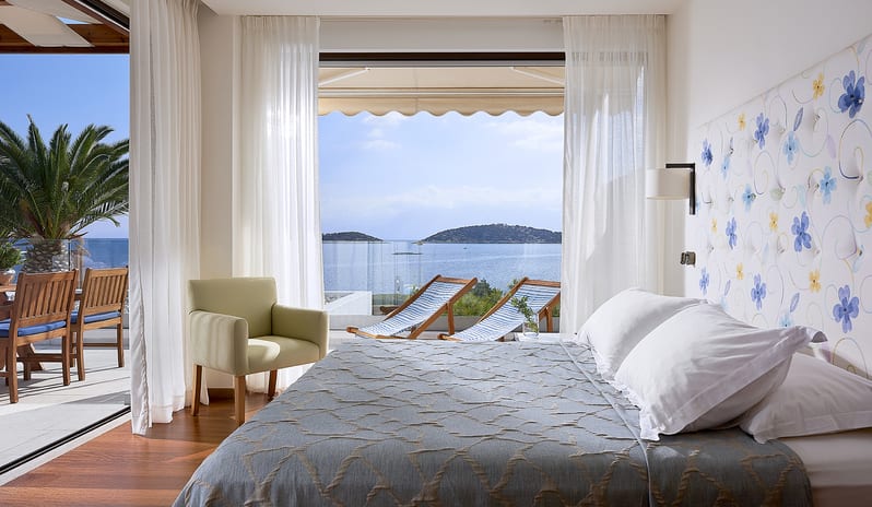 Club Suite One Bedroom Private Pool Sea View bedroom suite with an oceanfront terrace, blue floral accents, and a view of distant islands at St.Nicolas Bay Resort by Kenwood Travel