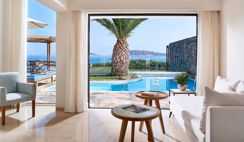 Club Suite One Bedroom Private Pool Sea View lounge with white sofas and stone flooring, offering a direct view of a private pool and a palm tree against a coastal backdrop at St.Nicolas Bay Resort by Kenwood Travel