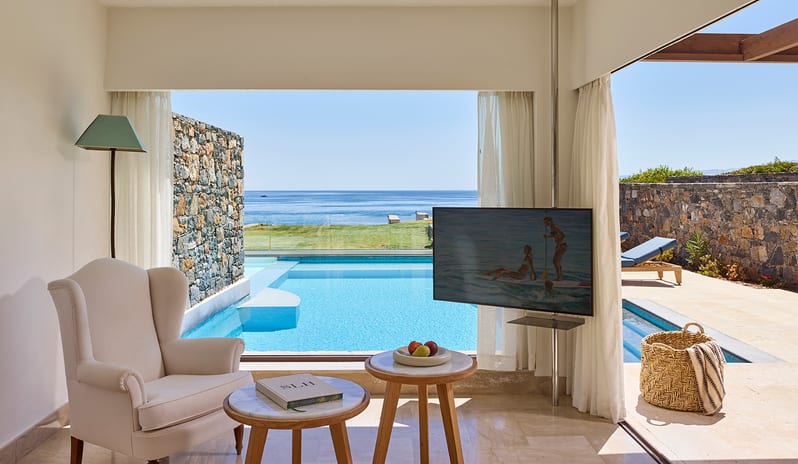 Club Suite One Bedroom Private Pool Sea View white wingback chair and marble side tables in a luxury suite, positioned in front of a floor-to-ceiling window showcasing a private infinity pool and the sea at St.Nicolas Bay Resort by Kenwood Travel